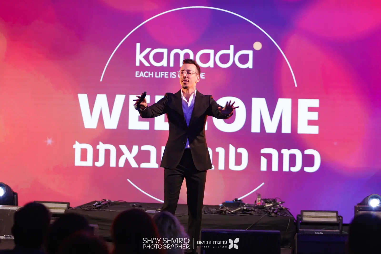 בן קלה מנחה כנס, מסך WELCOME מאחוריו